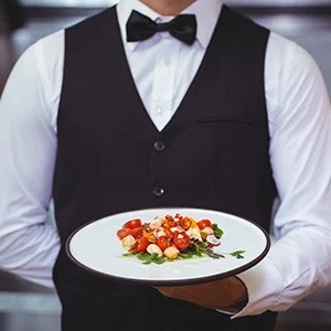 گارسون با لباس مشکی که بشقاب سفیدی با سبزیجات در دست دارد